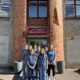 Gruppe von neun Schülern in blauen T-Shirts vor dem Eingang der Schule Sandoros mit einer litauischen Flagge.